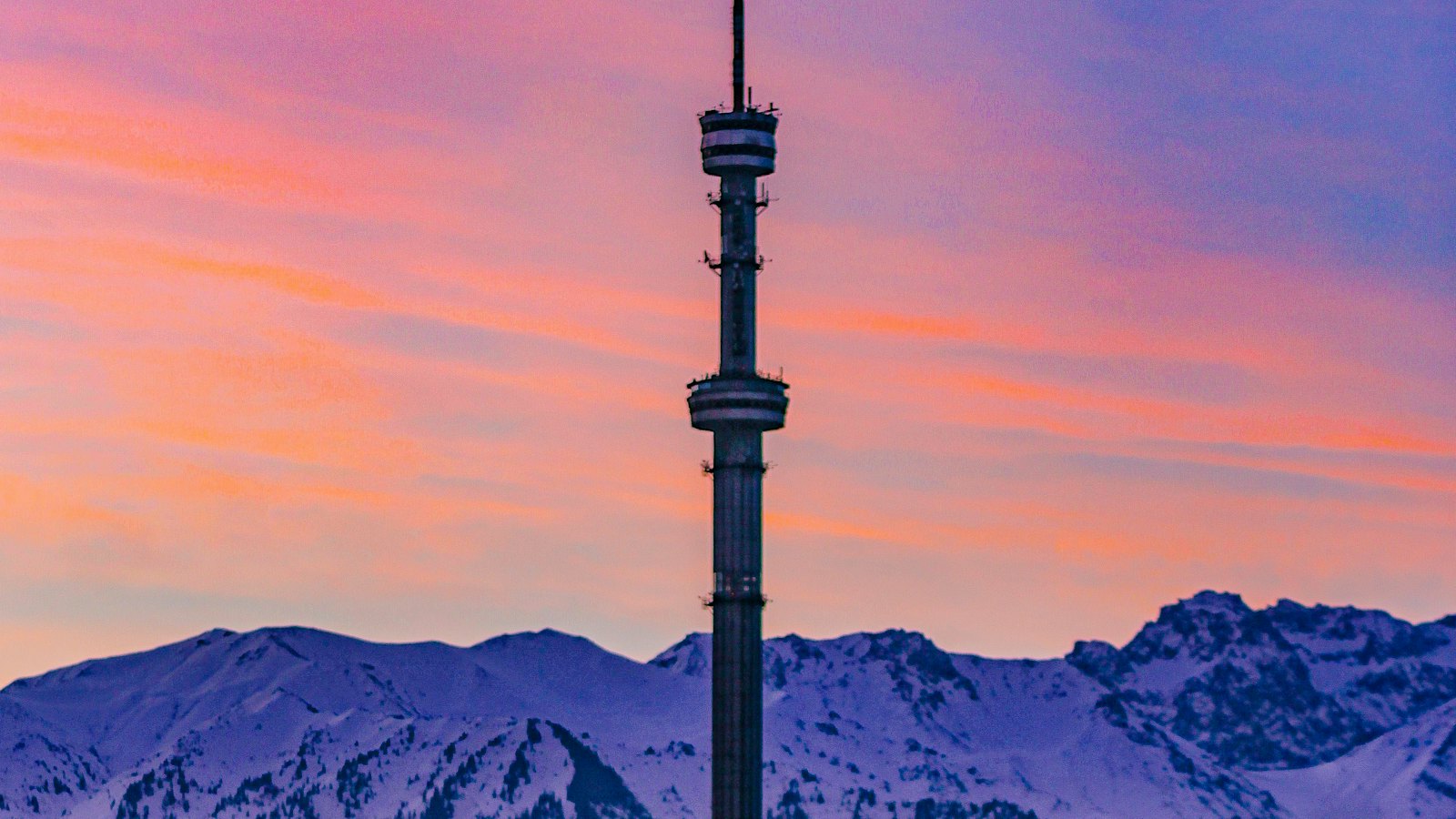 Almaty's Kok Tobe TV tower against a pink dusk sky and snowy Trans-Ili Alatau mountains