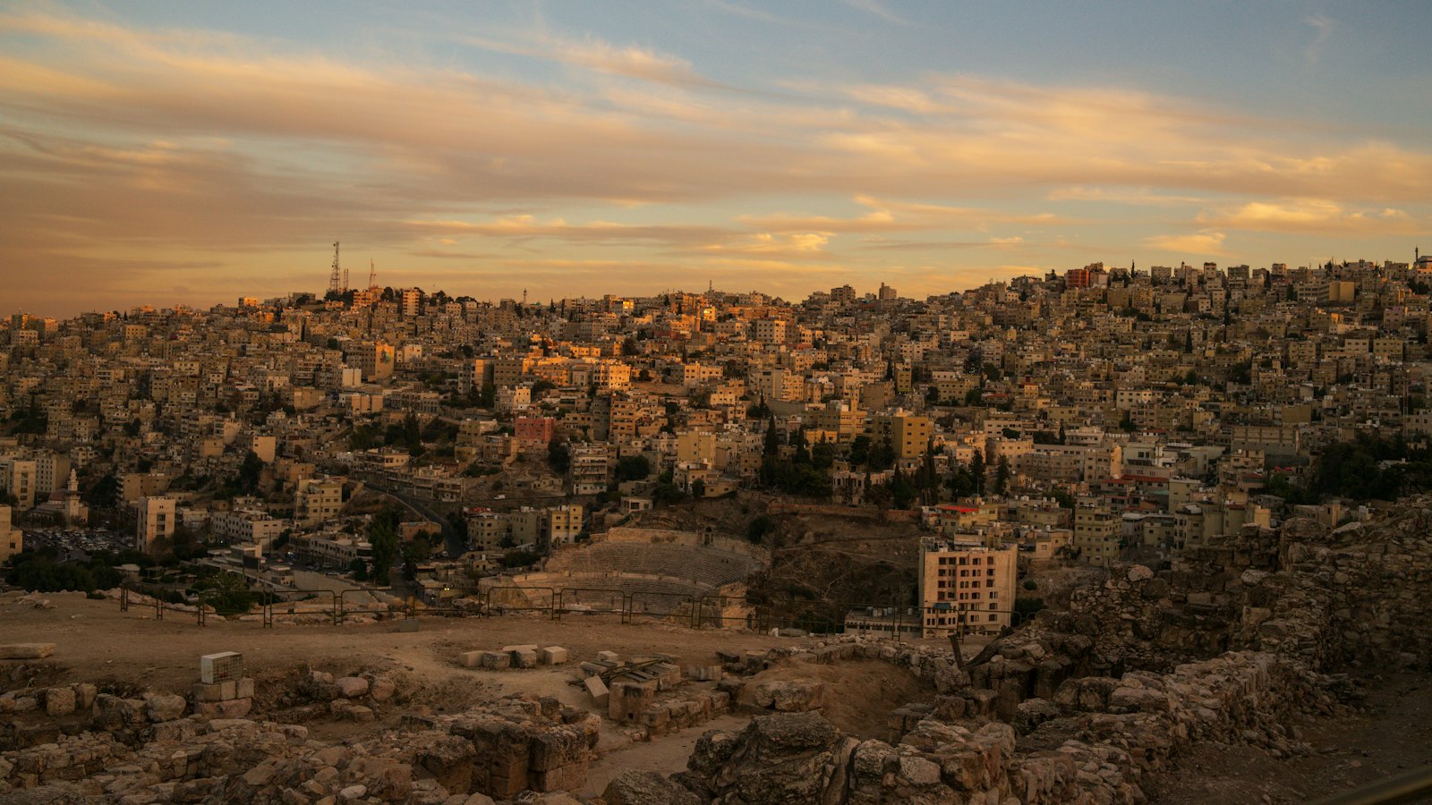 Amman's hillside houses and the Roman Theatre glowing under a golden dusk sky, seen from the Citadel