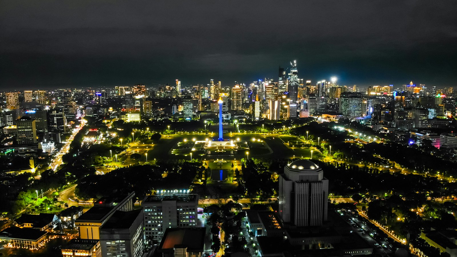 Aerial night view of Jakarta's blue-lit National Monument in Merdeka Square with the glowing city skyline behind