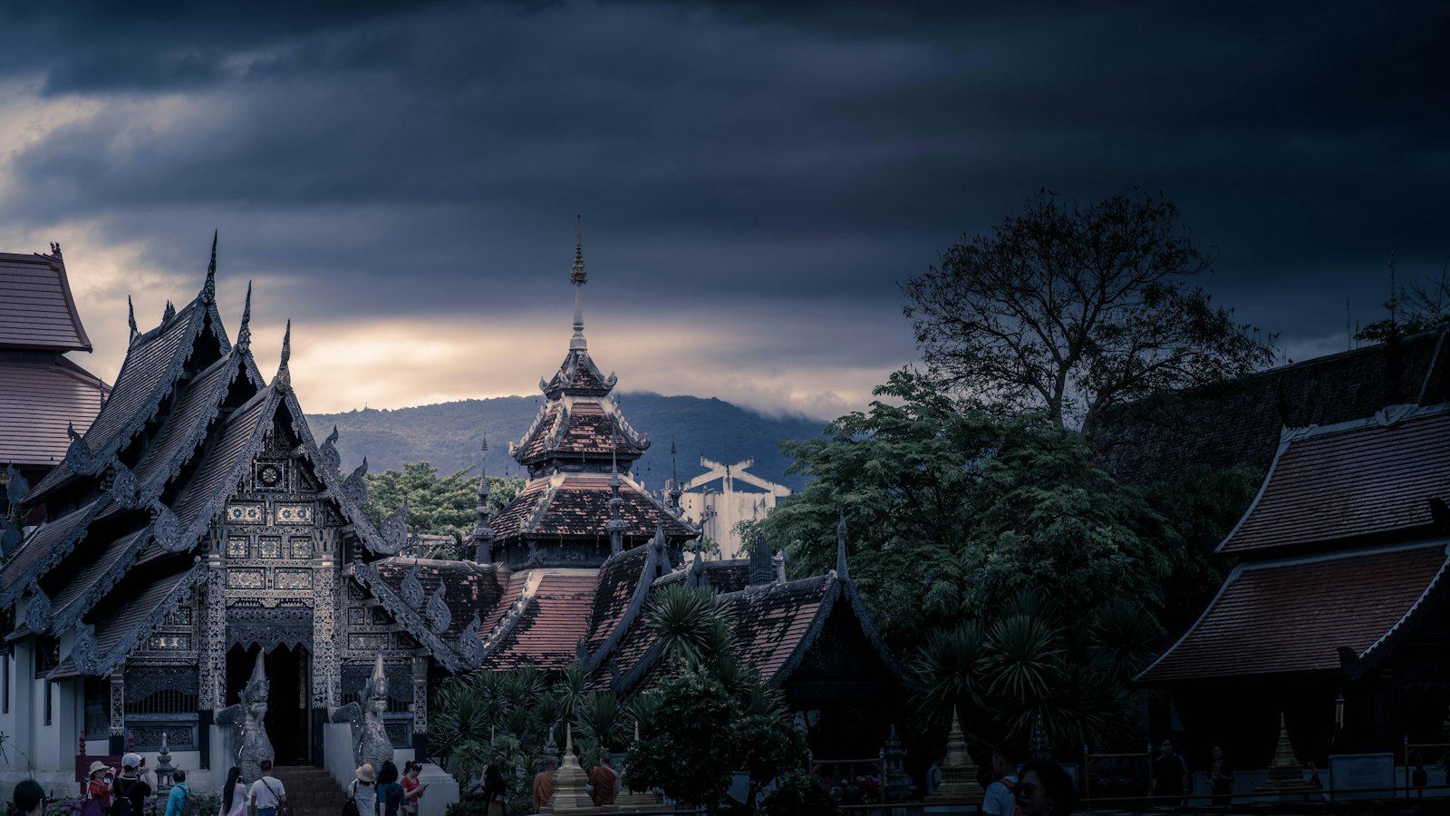 Ornate Lanna temple roofs in Chiang Mai beneath dark storm clouds and a glowing dusk horizon