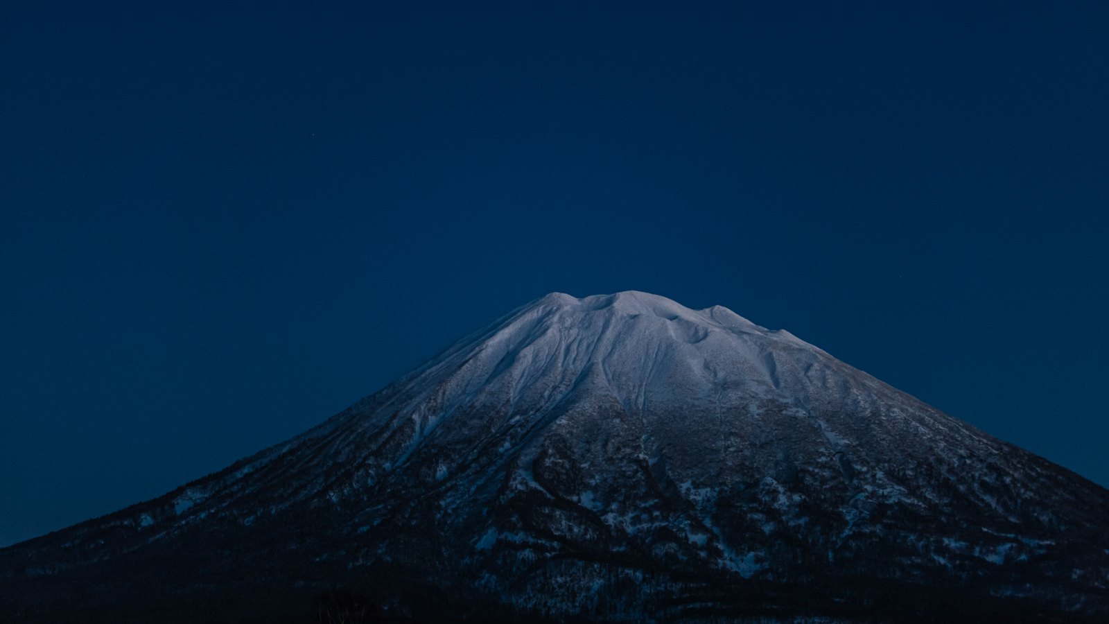 The conical snow-covered peak of Mount Yotei glowing under a deep blue night sky near Niseko