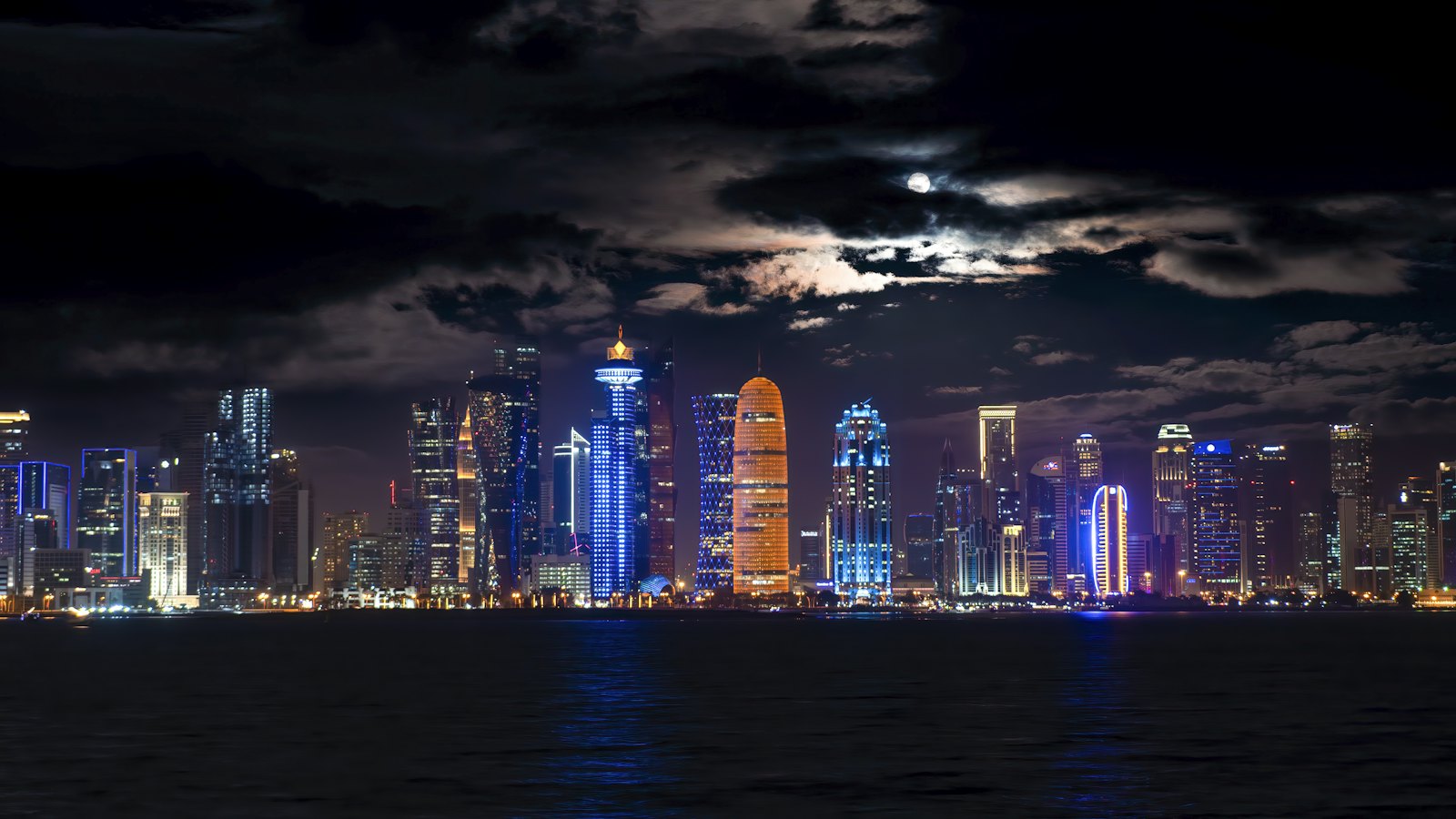 Doha's West Bay skyline glowing at night under moonlit clouds, seen across the bay