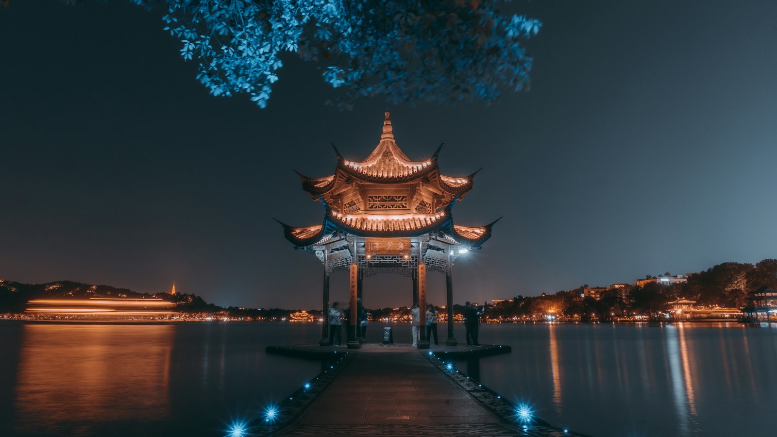 Jixian Pavilion lit up at night on West Lake, Hangzhou, with a stone causeway and city lights