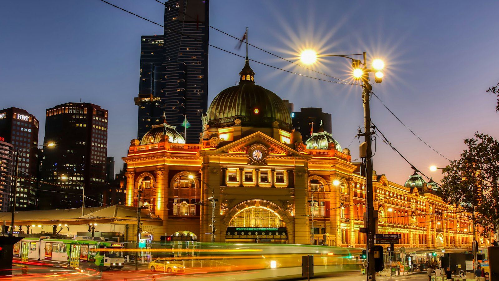 Flinders Street Station glowing gold at blue hour in Melbourne, with a tram streaking past