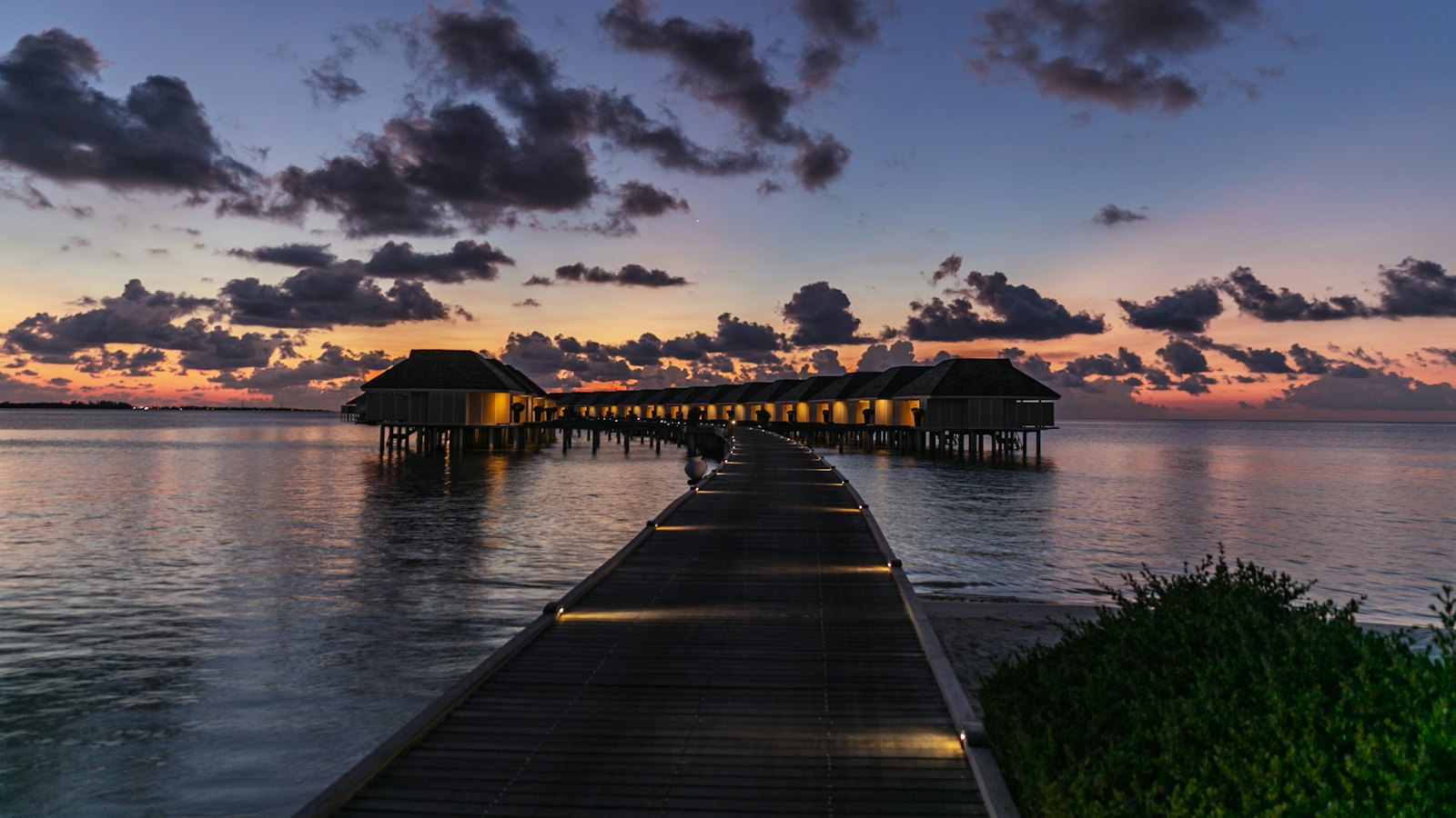 Lit wooden jetty leading to overwater villas at blue hour in South Ari Atoll, Maldives