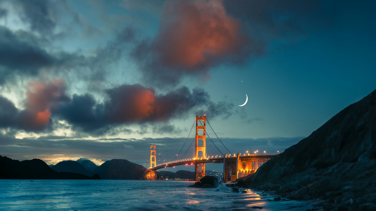 The Golden Gate Bridge glowing at dusk with a crescent moon over the bay