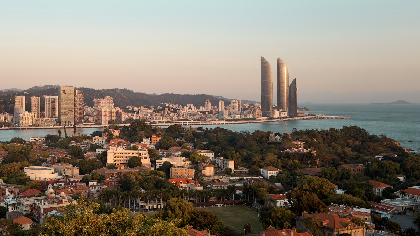 Shimao Straits Towers rising over Xiamen harbour in golden-hour light, seen above Gulangyu's rooftops