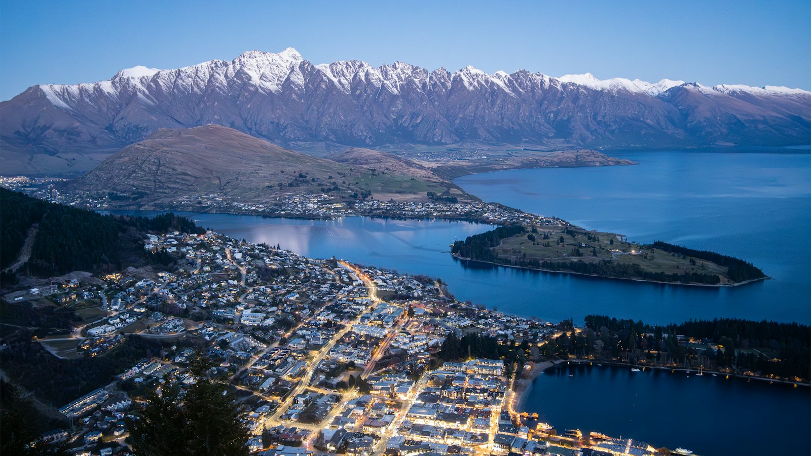 Queenstown's lights glowing beside Lake Wakatipu below the snow-capped Remarkables at blue hour